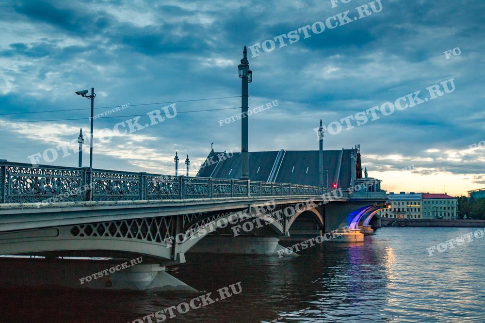 Композиция изображения акцентирует внимание на изяществе мостовой конструкции и архитектурных деталях Благовещенского моста в Петербурге, отраженных в водной глади. Этот снимок хорошо подойдет для туристических публикаций, посвященных архитектурному наследию города, или как элемент визуальной культуры в экспозициях, связанных с Петербургом.